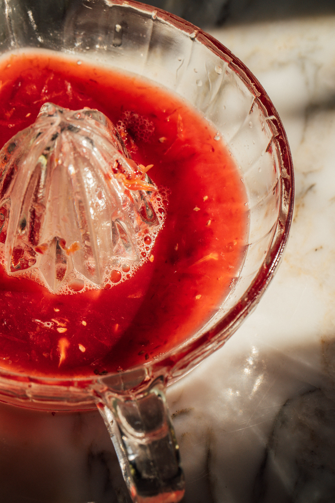 Fresh blood orange juice being pressed in a glass citrus juicer, with vivid red juice pooling around the reamer.